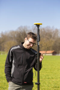 Im Vordergrund des Bildes steht ein Mitarbeiter des Projekts mit einem GNSS Vermessungsstab auf einer Wiese. Im Hintergrund ist ein Gebäude und Bäume zu sehen.