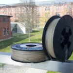 zwei Rollen auf denen durchsichtige runde Gummibänder aufgewickelt sind, im Hintergrund die Gebäude der Hochschule Ansbach