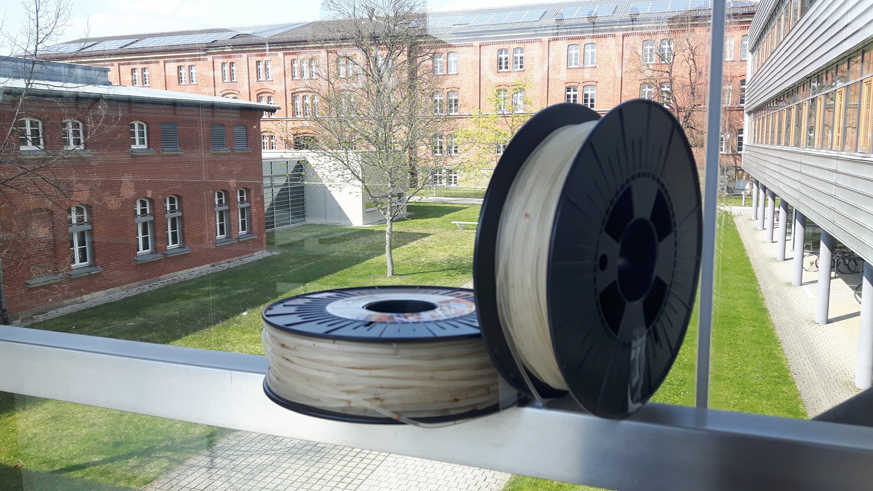 zwei Rollen auf denen durchsichtige runde Gummibänder aufgewickelt sind, im Hintergrund die Gebäude der Hochschule Ansbach