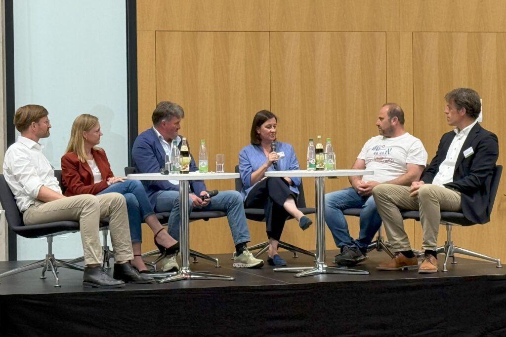 auf einem Podium in einem Hörsaal sitzen um zwei Tische gruppiert vier Männer und zwei Frauen