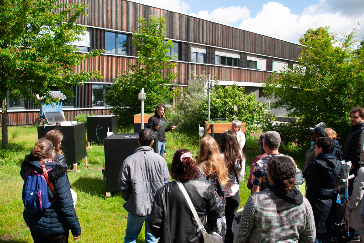 Unser Foto zeigt den BIT-Mitarbeitenden Matthias Wörlein steht vor Versuchsständen auf dem Campus Triesdorf und spricht zum Publikum. Beim Rundgang über den Campus informierten sich die Gäste zu den Themen Nachhaltigkeit und Ökologie. Unter anderem präsentierte der wissenschaftliche Mitarbeiter und Alumnus Matthias Wörlein das Forschungsprojekt Climate4Roofs. © Mechthild Müller