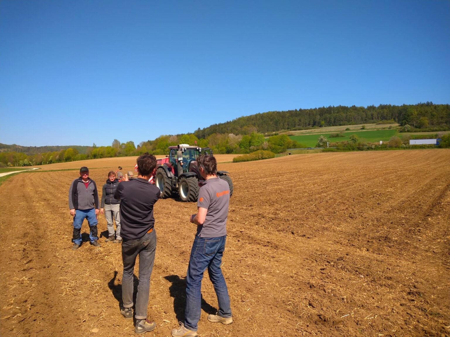 Ein Traktor mit einem Anhänger steht auf einem Acker. Daneben stehen 2 Personen, die man von vorne sieht und 2 Personen mit dem Rücken zur Kamera.