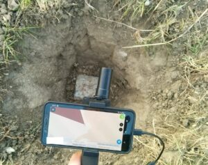 Auf dem Bild ist im Hintergrund ein Grenzstein im Boden zu sehen. Im Vordergrund ist das Display des GNSS Vermessungsstabes zu sehen.