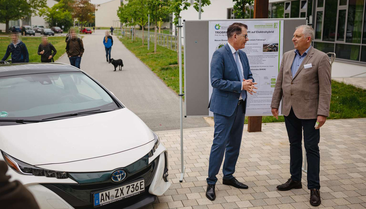 Dr. Gerd Müller mit Prof. Norbert Huber unterhalten sich neben dem Triobrid, dahinter ein Plakat zum Triobrid